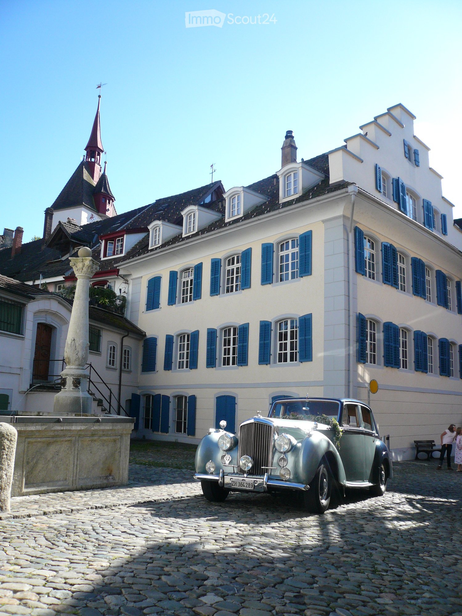 A large, multi-story building with white walls and blue shutters, featuring a classic car parked in front on a cobblestone street.