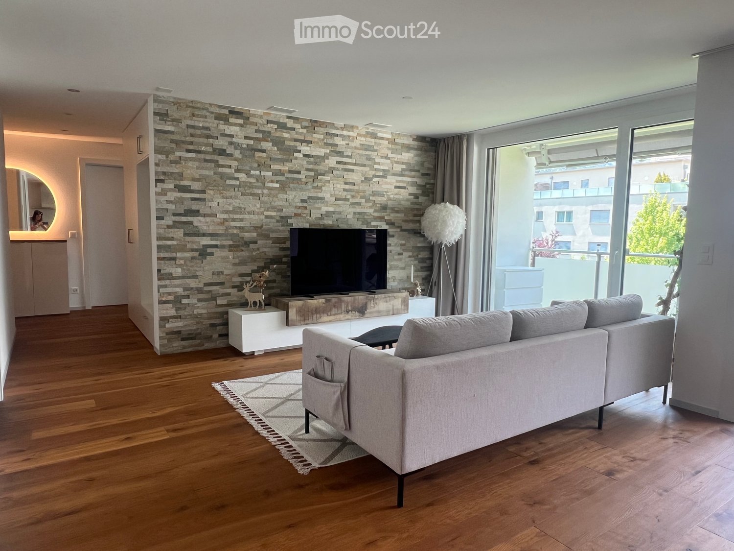 Modern living room with wooden flooring, a grey sectional sofa, a coffee table, a television on a white stand, a floor lamp, and a decorative stone wall.