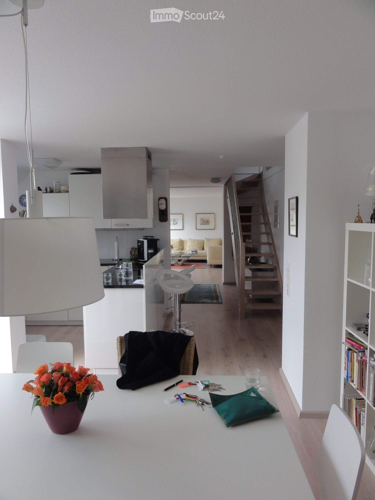 Open-plan living and dining area with a white dining table, chairs, a bar stool, a sofa in the background, a bookshelf, and a kitchen area with cabinets and appliances.