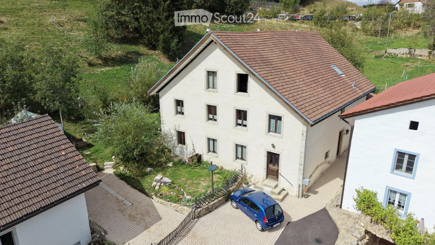Aerial view of a two-story house with a red-tiled roof, multiple windows, and a small garden area. A blue car is parked in the driveway.