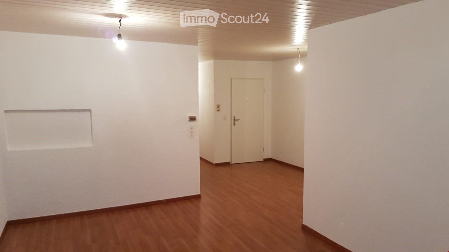 Empty room with wooden flooring, white walls, and a single door leading to another space. The room has recessed lighting and a small window on one wall.