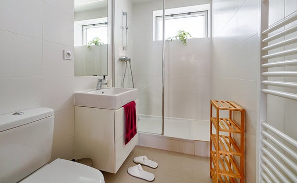 Modern bathroom with white tiles, a sink, mirror, toilet, glass-enclosed shower, a red towel, a wooden shelf, and white bathroom slippers on the floor.
