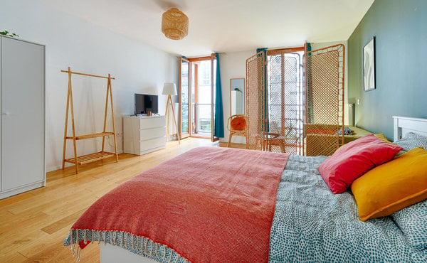 Bright bedroom with wooden floor, large windows, balcony access, bed with colorful blankets, dresser, chair, and screen.