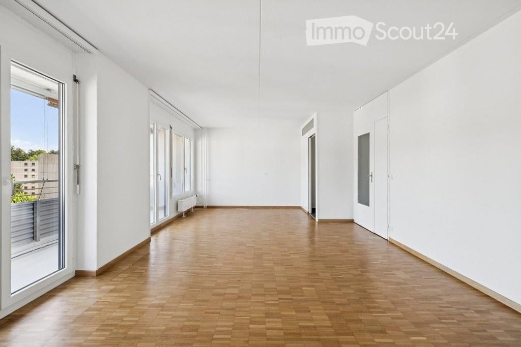 Bright, empty room with wooden flooring, white walls, large windows, and a balcony access point.
