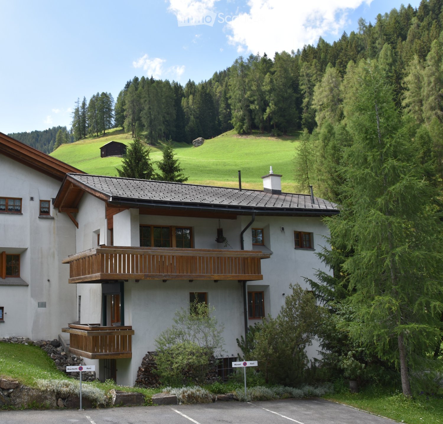 Two-story white house with wooden balconies, surrounded by trees and a grassy hill in the background.