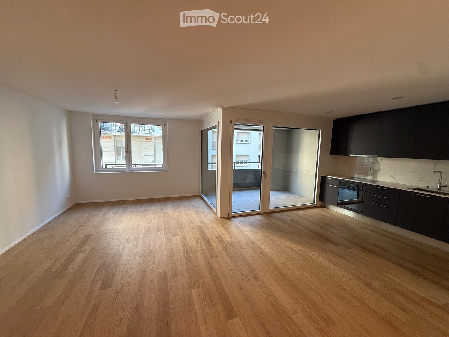 Empty room with wooden flooring, large windows, and a small kitchen area with cabinets and a sink.
