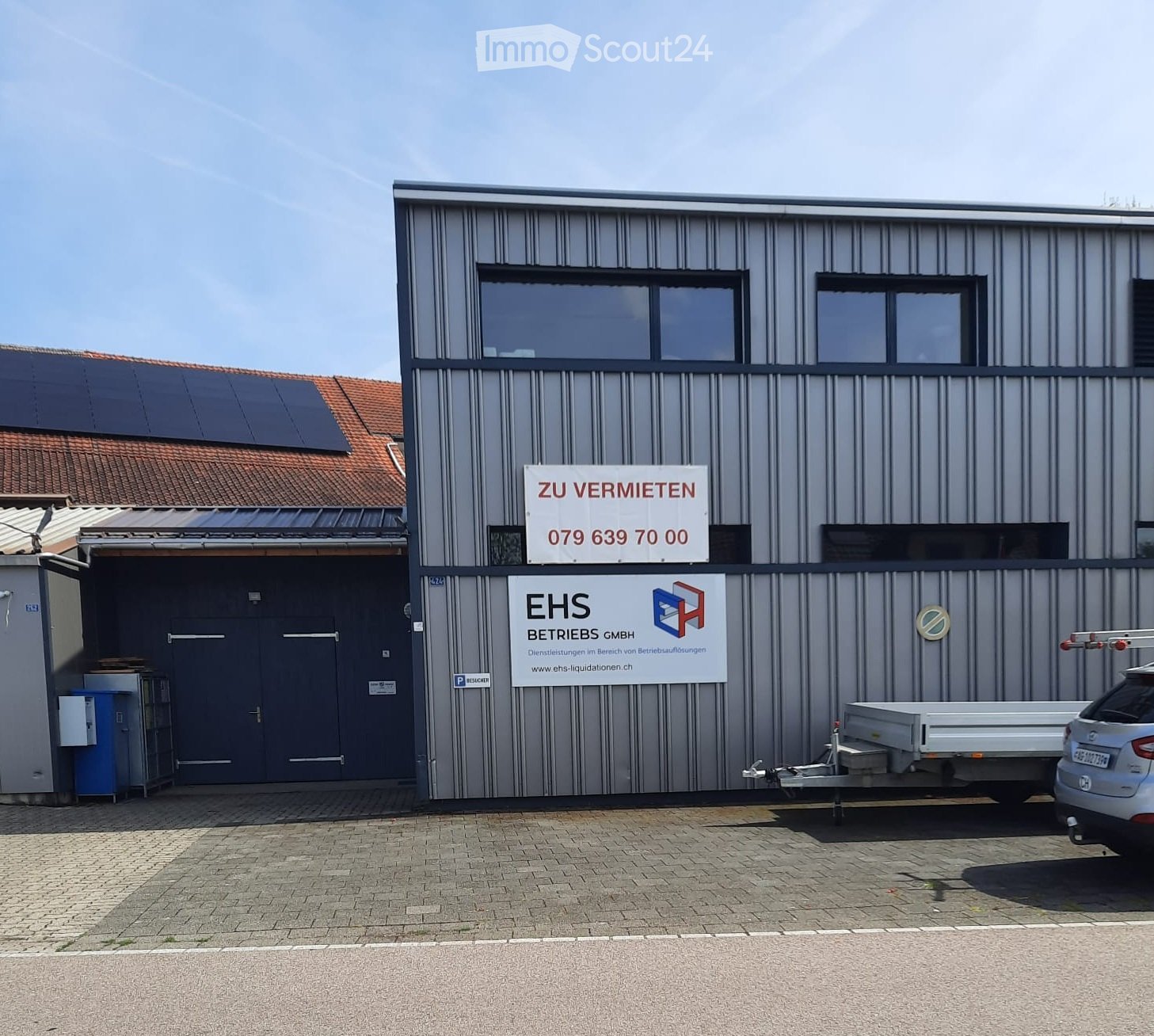 Modern industrial building with grey metal siding, large windows, and signage reading "ZU VERMIETEN" and "EHS BETRIEBS GMBH".