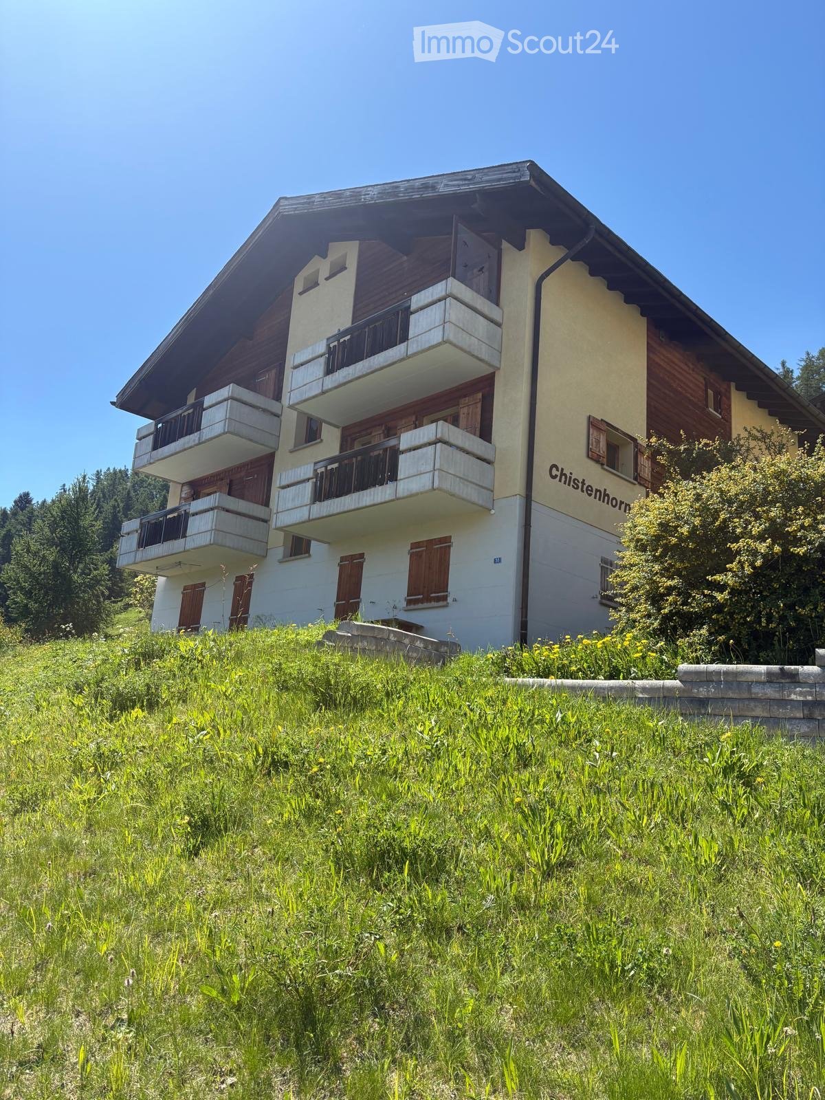 Two-story house with balconies, wooden shutters, and a grassy slope in front.