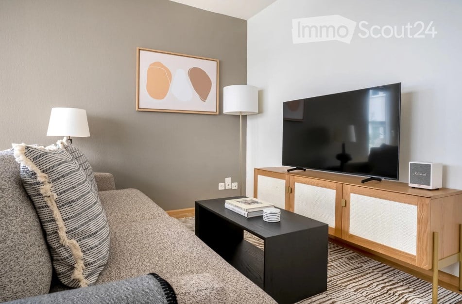 Modern living room with a grey sofa, black coffee table, floor lamps, flat-screen TV on a wooden stand, and a decorative picture frame on the wall.