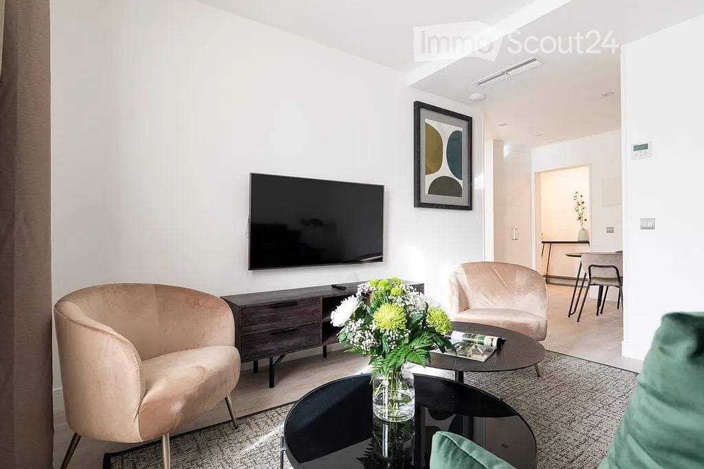 Modern living room with a sleek black television mounted on the wall, a dark wooden TV stand, a round coffee table with a flower arrangement, two light-colored armchairs, and a dining area visible in the background.