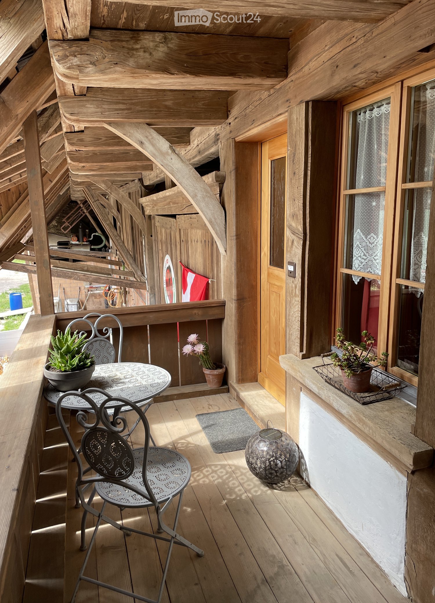 Wooden balcony with a table, two chairs, potted plants, a decorative sphere, and a small rug.