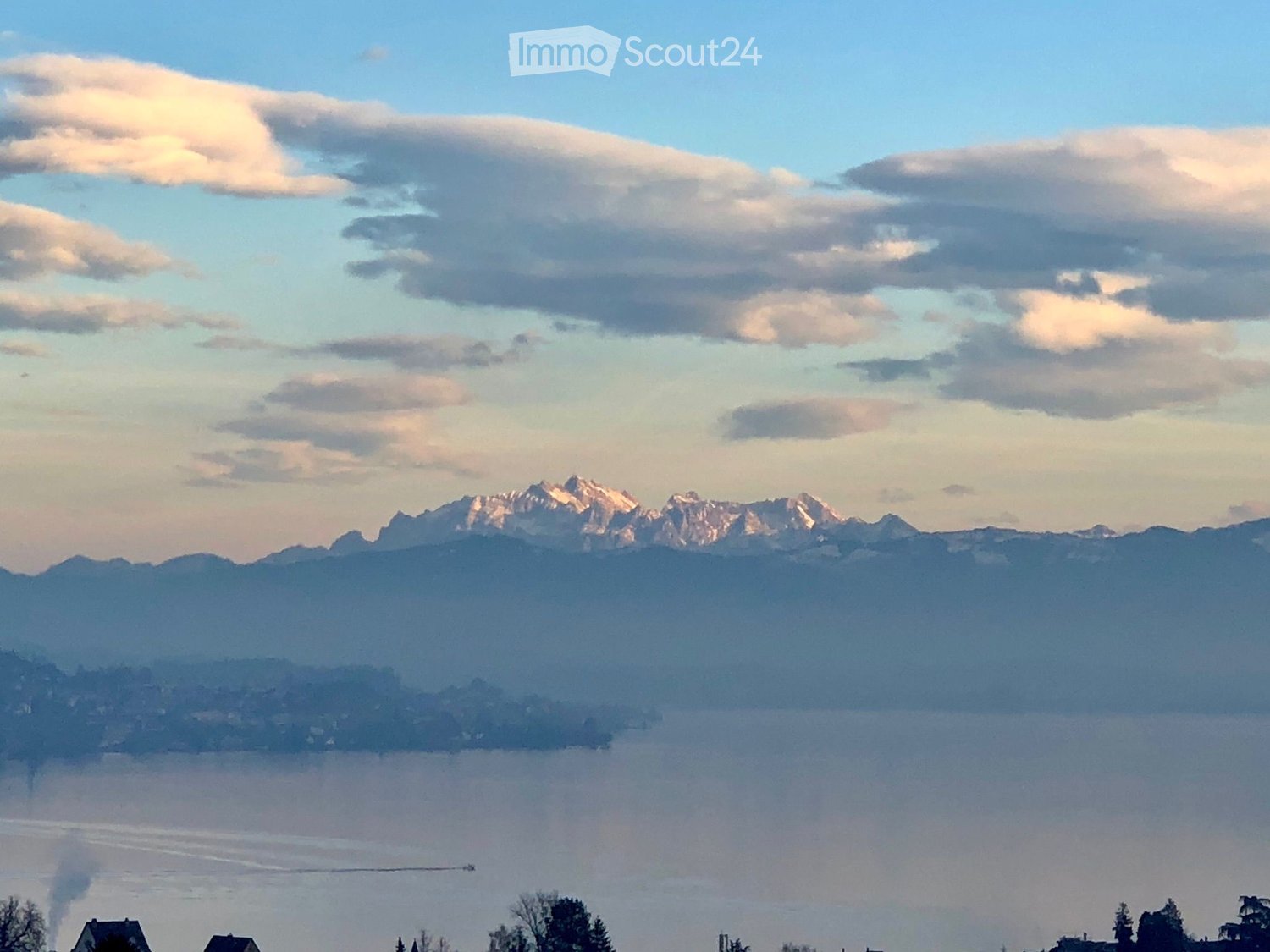 Panoramic view of a lake surrounded by mountains under a partly cloudy sky, with a few houses visible on the shore.