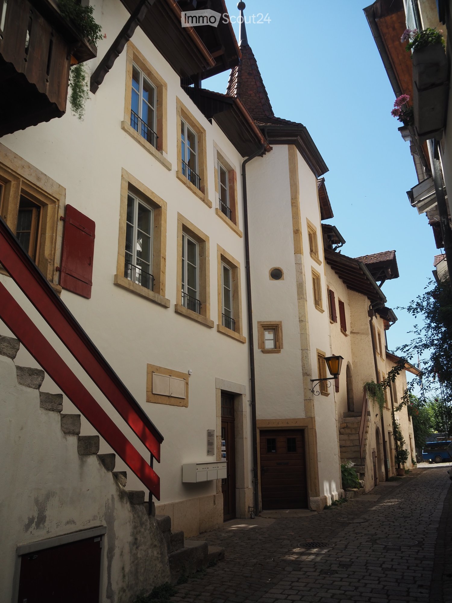 Narrow cobblestone street lined with traditional European buildings featuring white plaster walls, wooden shutters, and small balconies. The architecture includes a mix of multi-story structures with pitched roofs and decorative elements.