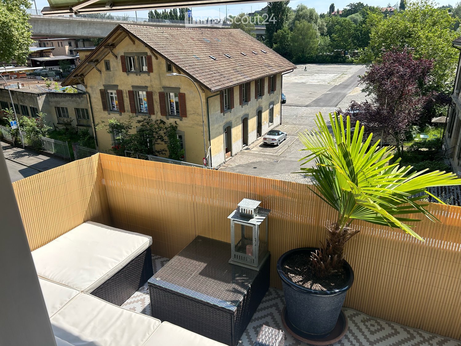 a yellow house in the background, a terrace with a couch, a table, a lantern, and a potted plant