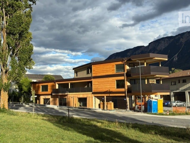 Modern multi-story building with wooden facade, balconies, and large windows, situated in a scenic area with mountains in the background.