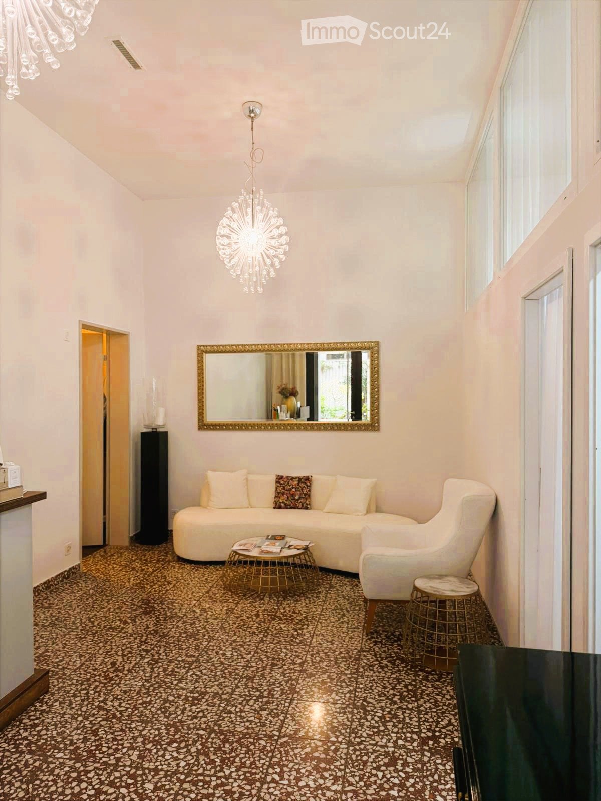 Living room with a white sofa, coffee table, armchair, and decorative mirror. The room features a patterned tile floor and a modern chandelier.