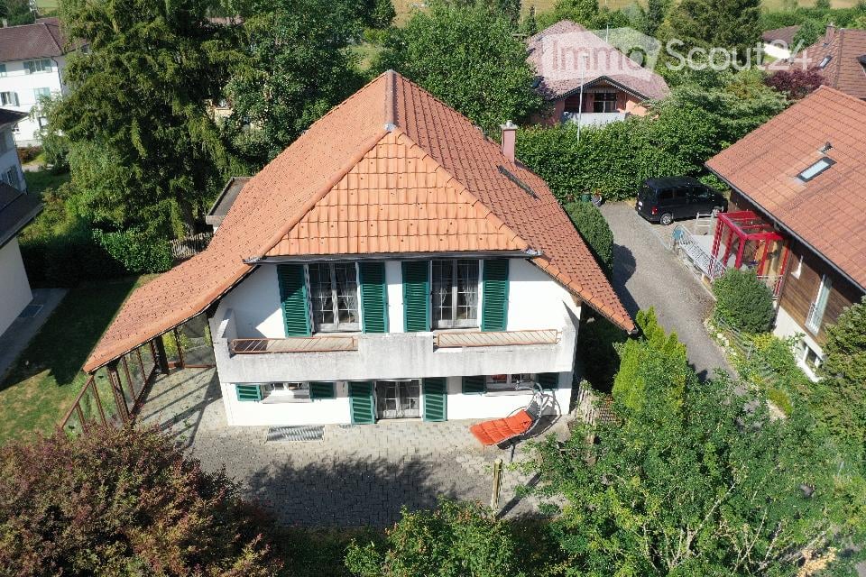 Aerial view of a two-story house with a red-tiled roof, white walls, green shutters, and multiple windows. The house has a covered patio area with outdoor furniture and is surrounded by greenery and neighboring houses.