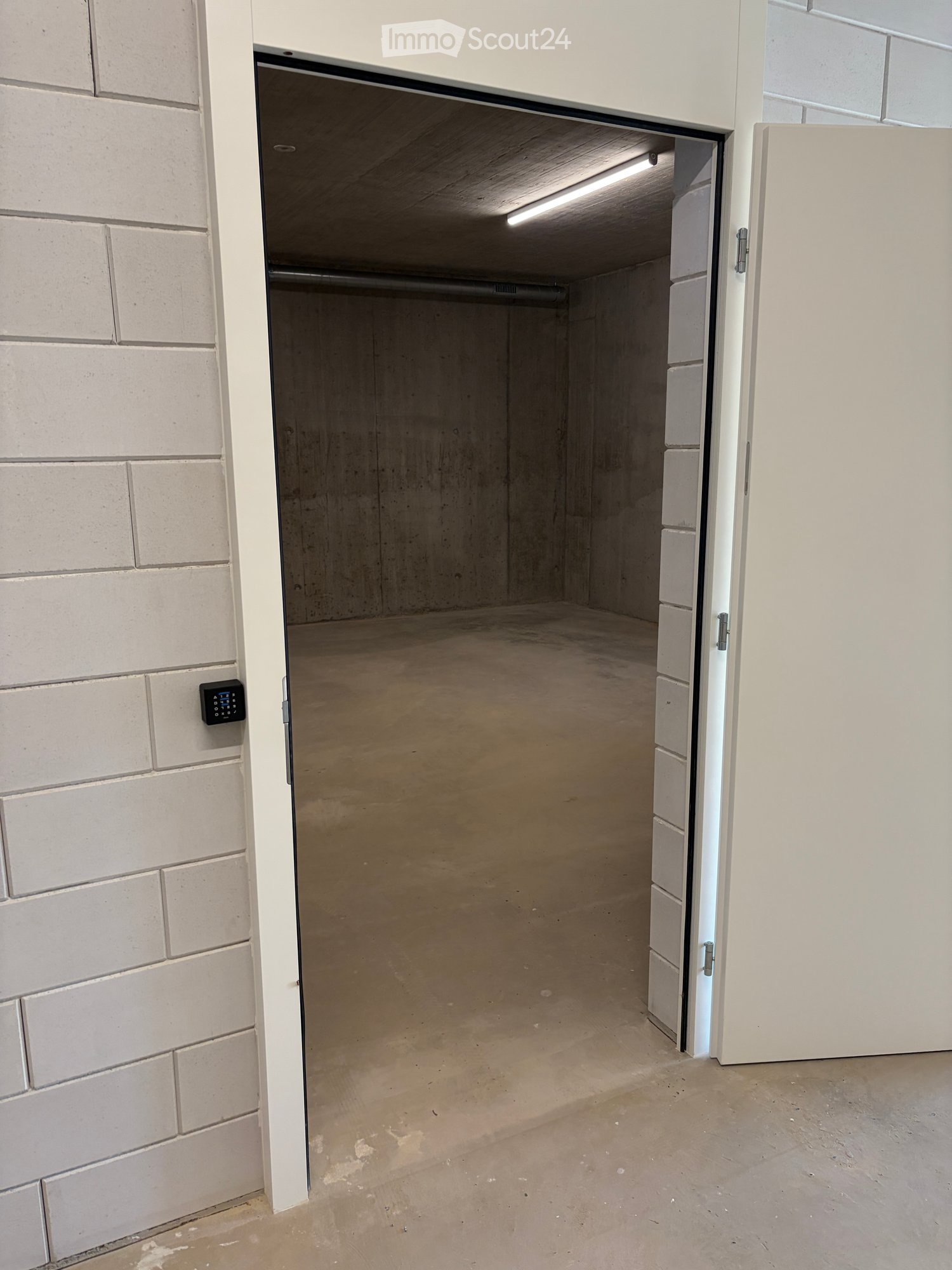 Empty garage with concrete floor, white brick walls, and an open door leading to a concrete-floored space.