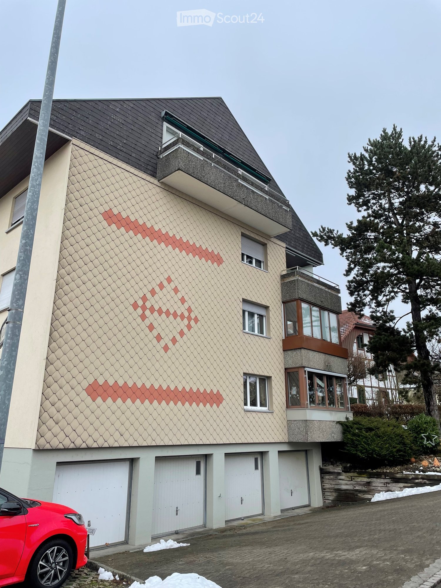 Two-story house with beige siding, red diamond-patterned accents, multiple windows, and a balcony on the upper level. Features a garage with three doors and a raised ground floor.