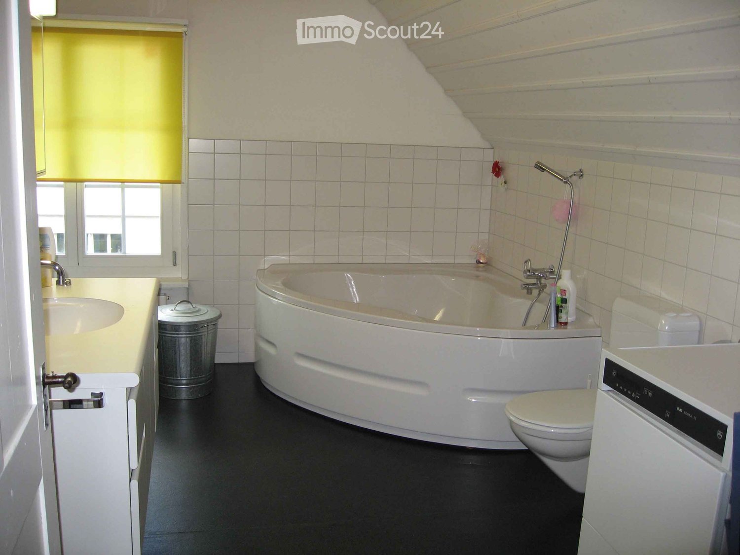 Bathroom with a bathtub, toilet, sink, and a window with blinds. The room features white tiled walls and a black floor.