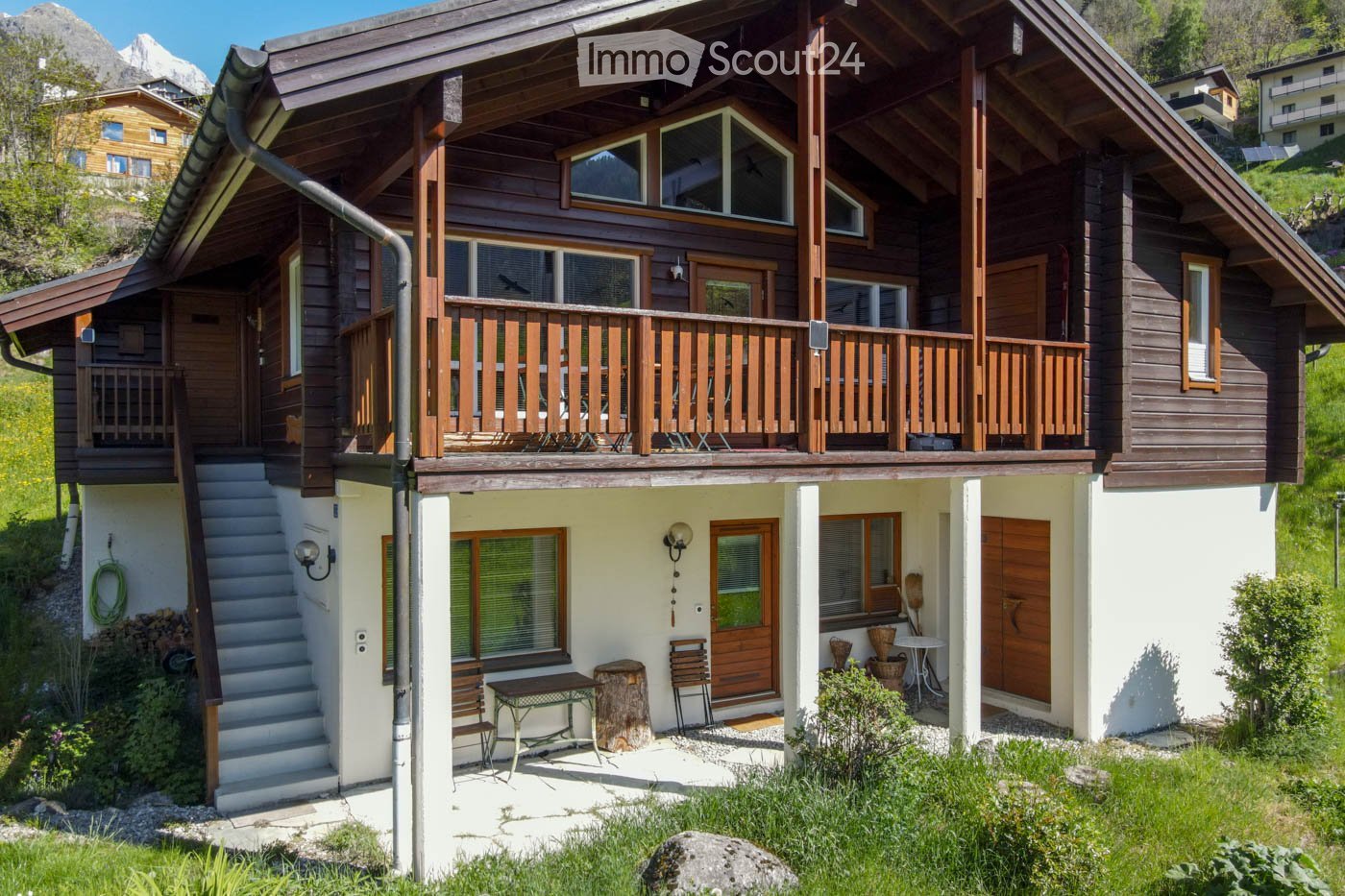 House. Two-story wooden house with a balcony, surrounded by greenery and mountains in the background.
