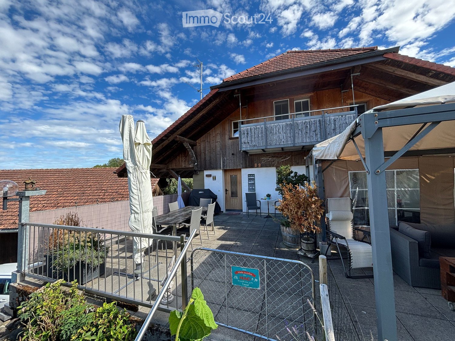 Outdoor patio area with a wooden table, chairs, a sofa, potted plants, and an umbrella. The patio is enclosed by a metal railing and overlooks a wooden house with a balcony.
