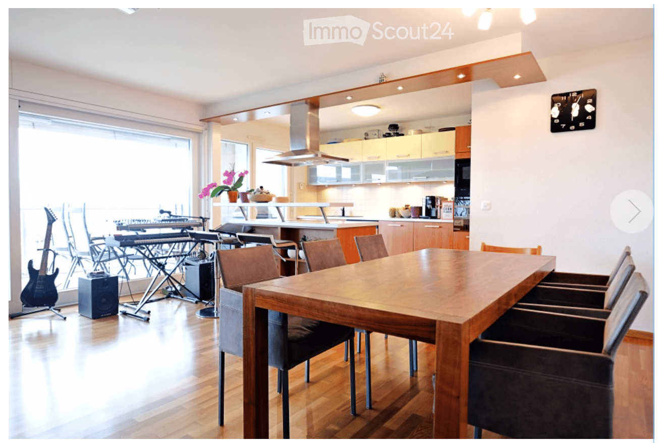 Open-plan living and dining area with a large wooden dining table, chairs, a guitar, keyboard, drum set, and a modern kitchen with cabinets, appliances, and a clock on the wall.