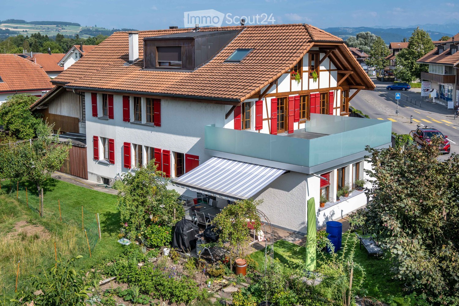A two-story house with red shutters and a tiled roof, featuring a modern balcony and a well-maintained garden with outdoor furniture.