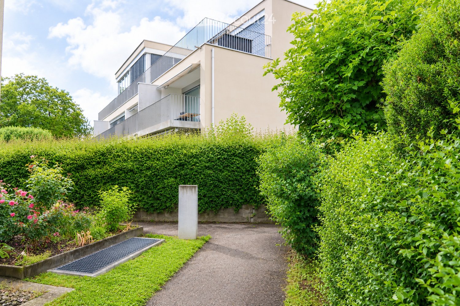 Modern residential building with raised ground floor, surrounded by lush greenery and a well-maintained garden path.