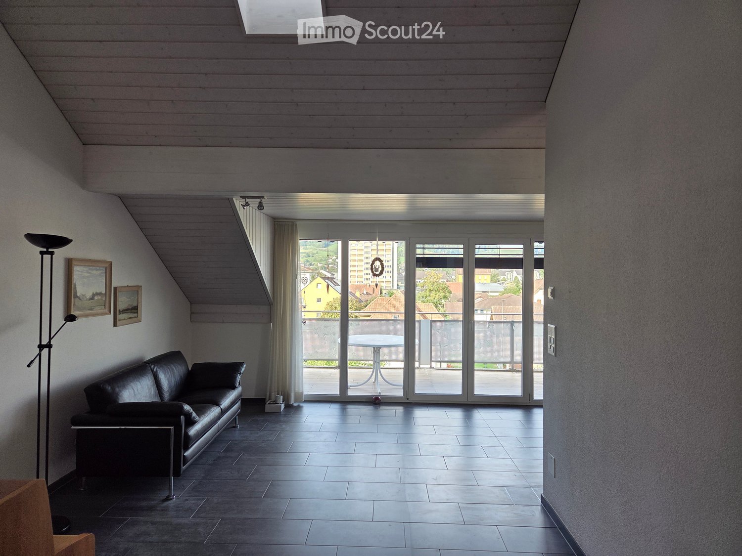 Modern living room with dark tiled floor, black leather sofa, floor lamp, and large sliding glass doors opening to a balcony with a view of the city.