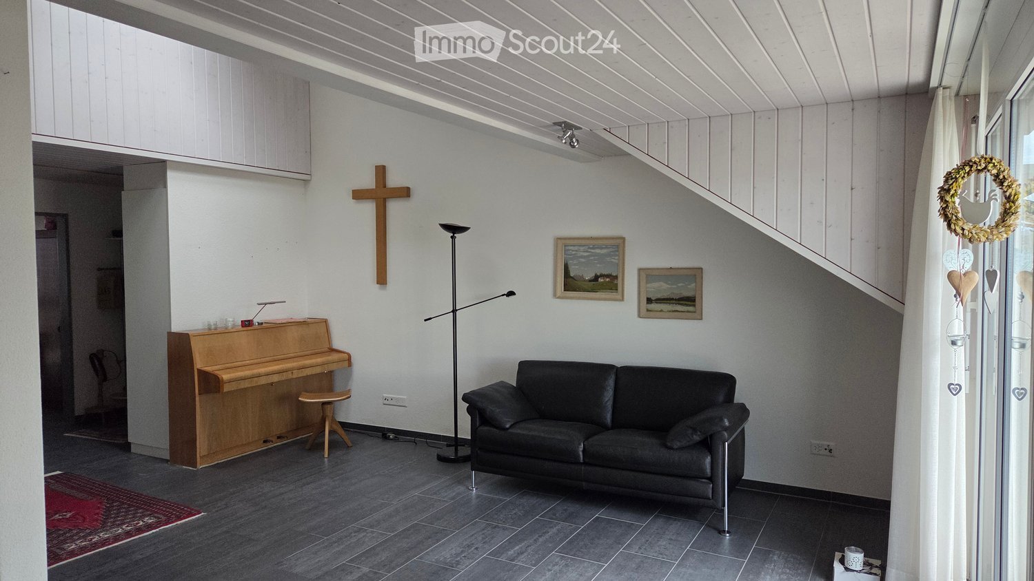 Living room with a dark grey sofa, wooden piano, floor lamp, cross on the wall, and two picture frames.