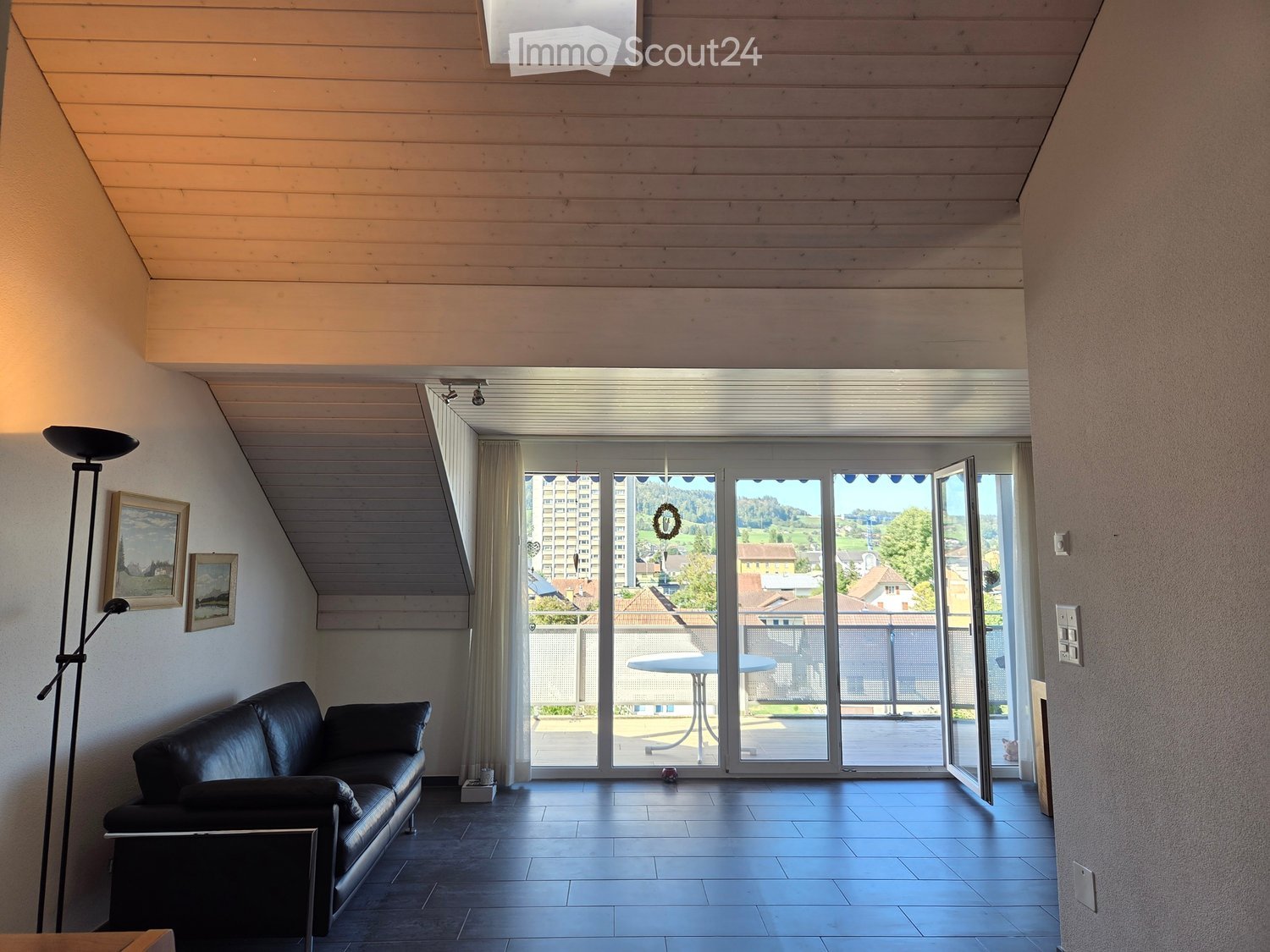 Modern living room with dark tiled floor, black leather sofa, floor lamp, picture frames, and large sliding glass doors opening to a balcony with outdoor furniture.