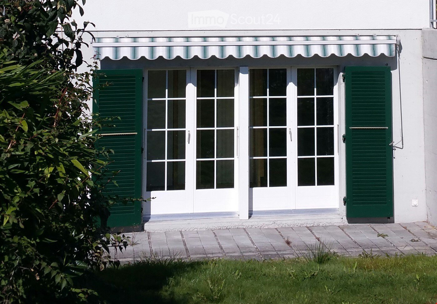 White building with green shutters, large windows, and a striped awning above the entrance.