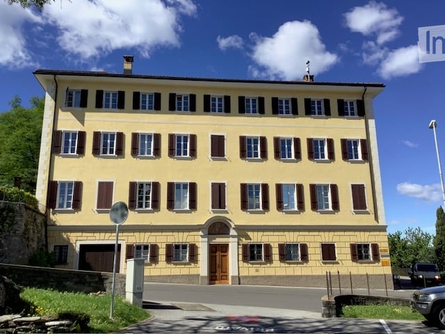 A large, yellow, multi-story building with numerous windows and brown shutters, featuring a central arched entrance and a small balcony above it.