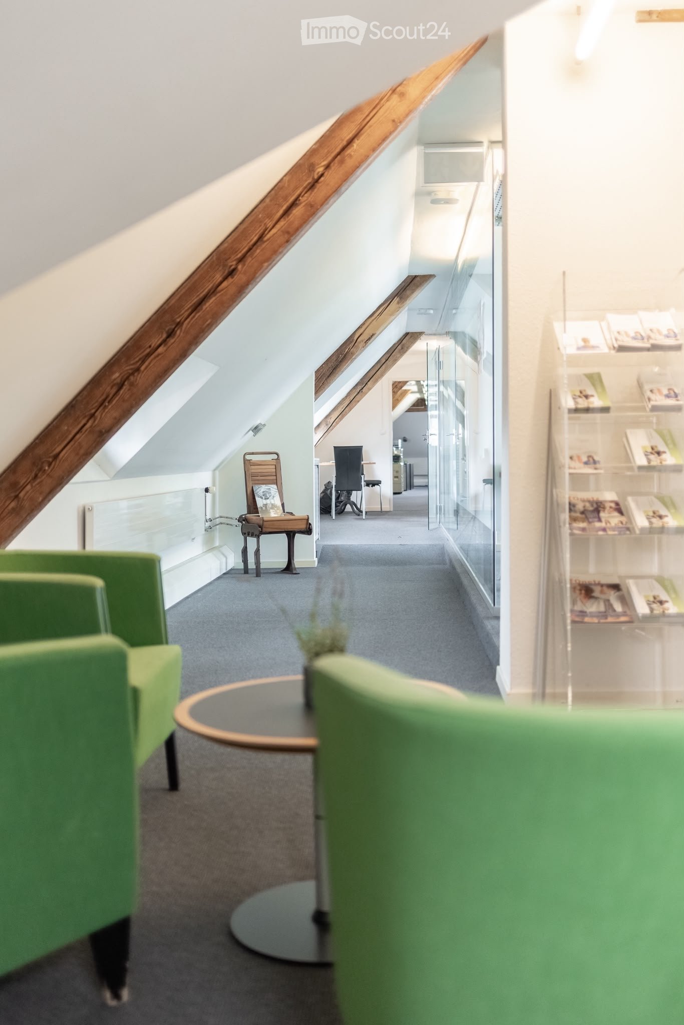 Modern hallway with green chairs, a round table, and a wooden chair with a picture frame on it. Shelves with books and magazines are visible in the background.