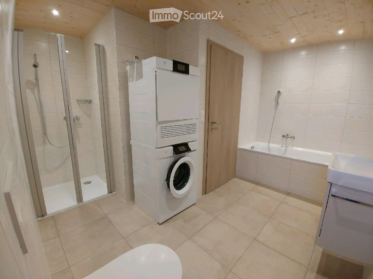 Modern bathroom with a white tiled shower, bathtub, toilet, and a stacked washing machine and dryer unit.