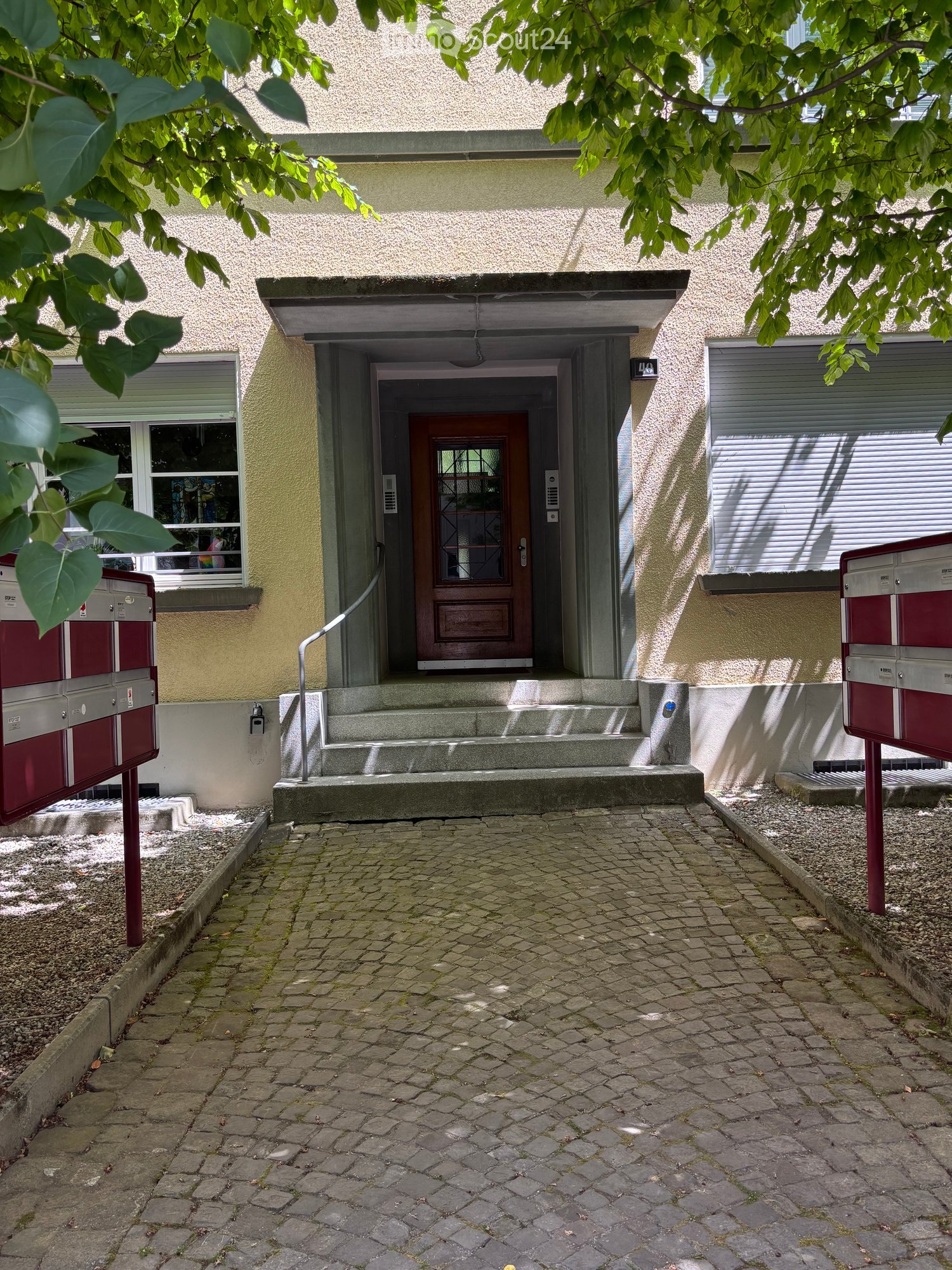 Entrance to a two-story building with a raised ground floor, featuring a wooden door, steps, and mailboxes on either side.