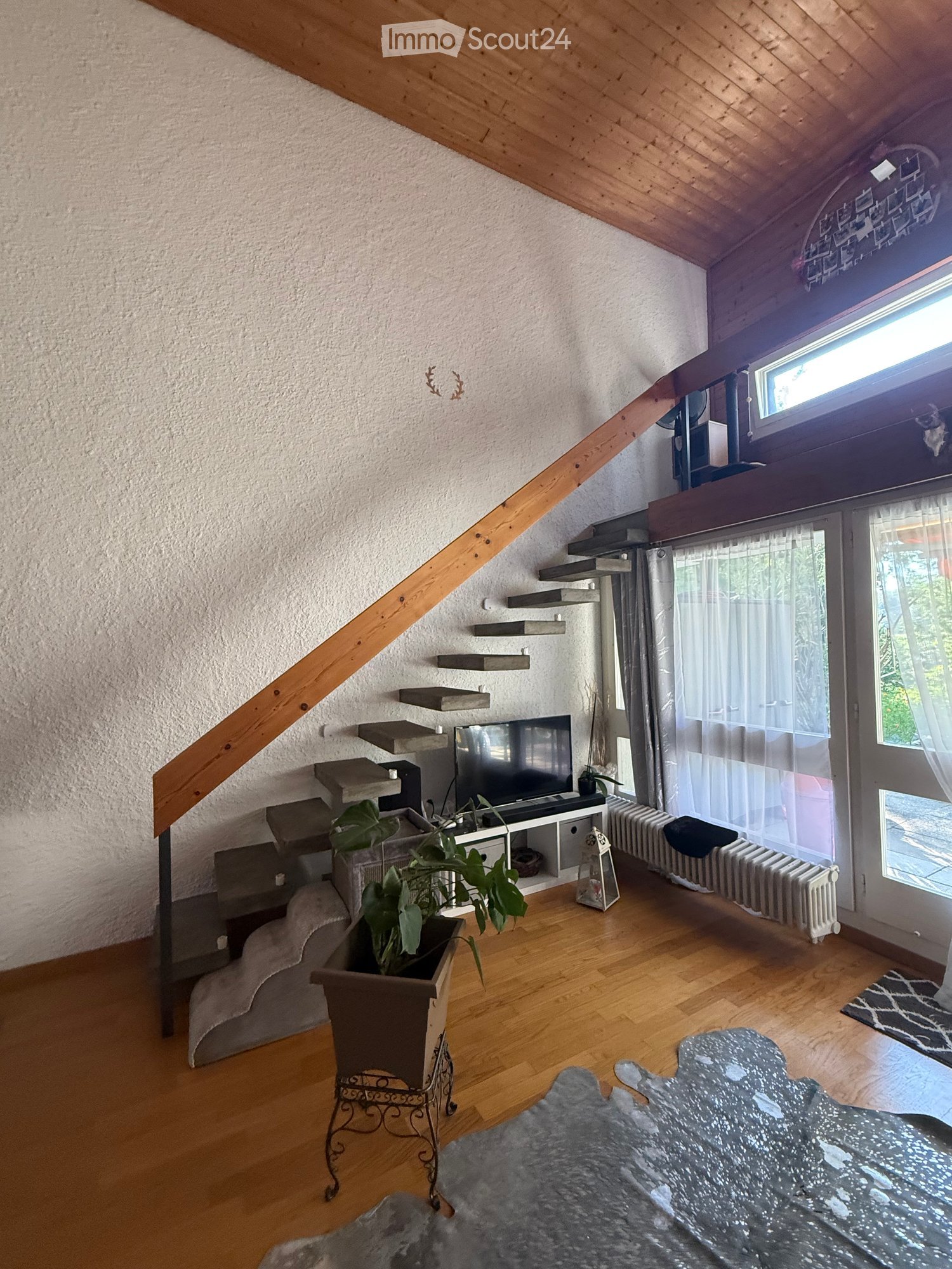 A cozy living room with wooden floors, a staircase leading upwards, a sofa, a coffee table, a television, and a potted plant. The room features a radiator and several shelves along the wall.