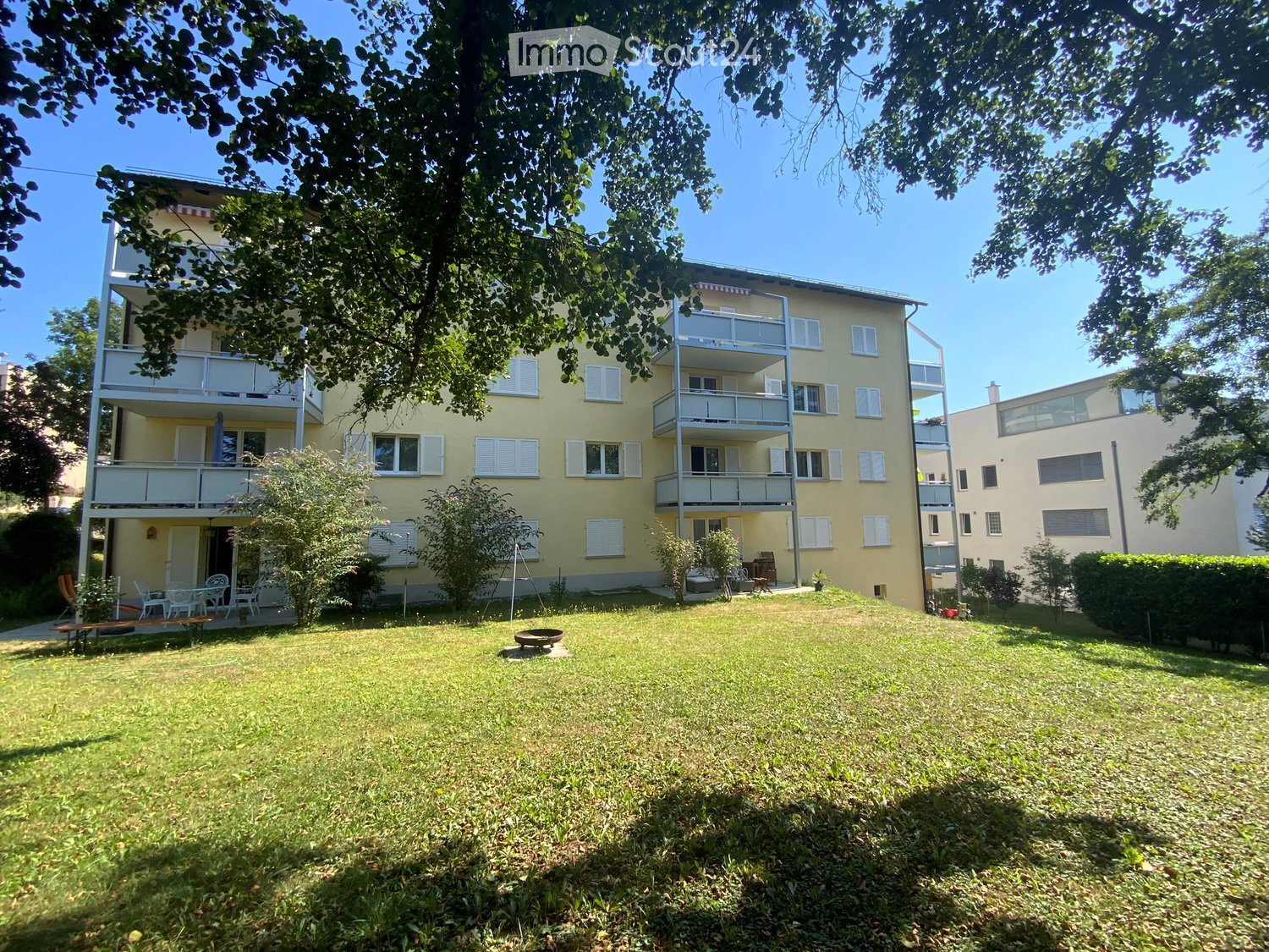 A multi-story yellow building with balconies, surrounded by a grassy yard with outdoor furniture and trees.