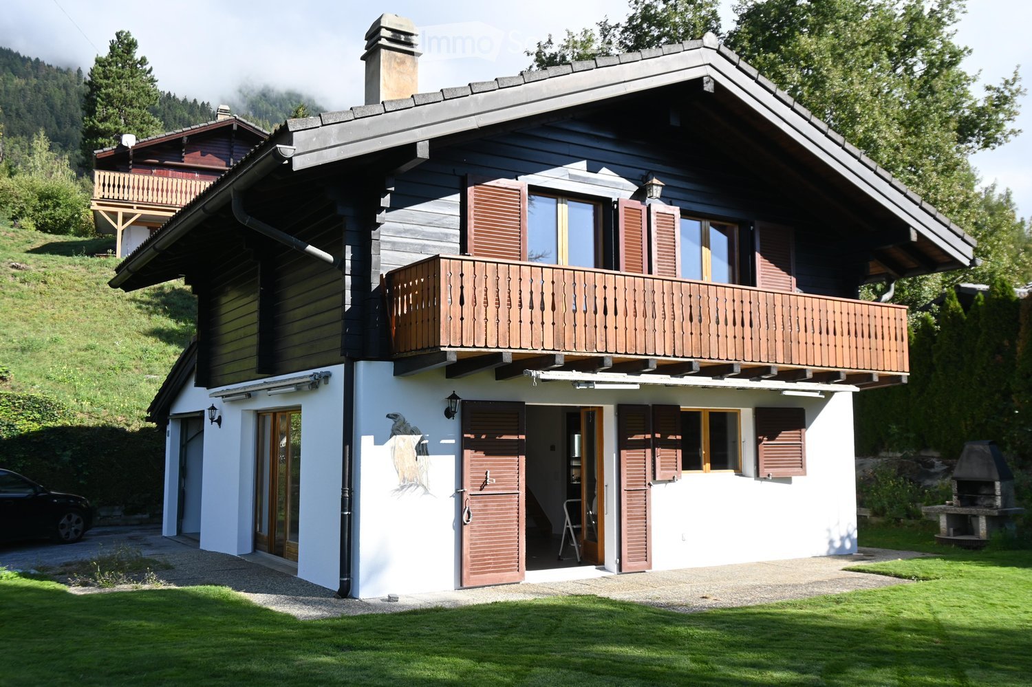 Two-story wooden house with a balcony, white lower facade, and dark upper facade. Features include a chimney, multiple windows with shutters, and a well-maintained lawn.