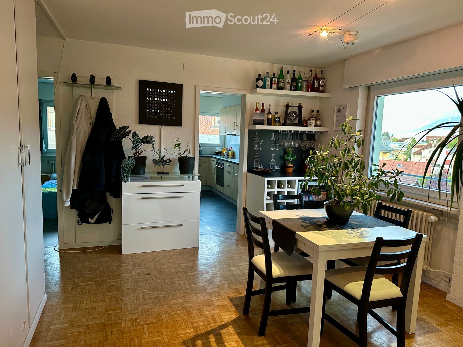 A modern, well-lit living and dining area with wooden flooring, a white dining table with four chairs, a potted plant, a white cabinet, and a view of a kitchen area with white cabinets and a refrigerator.