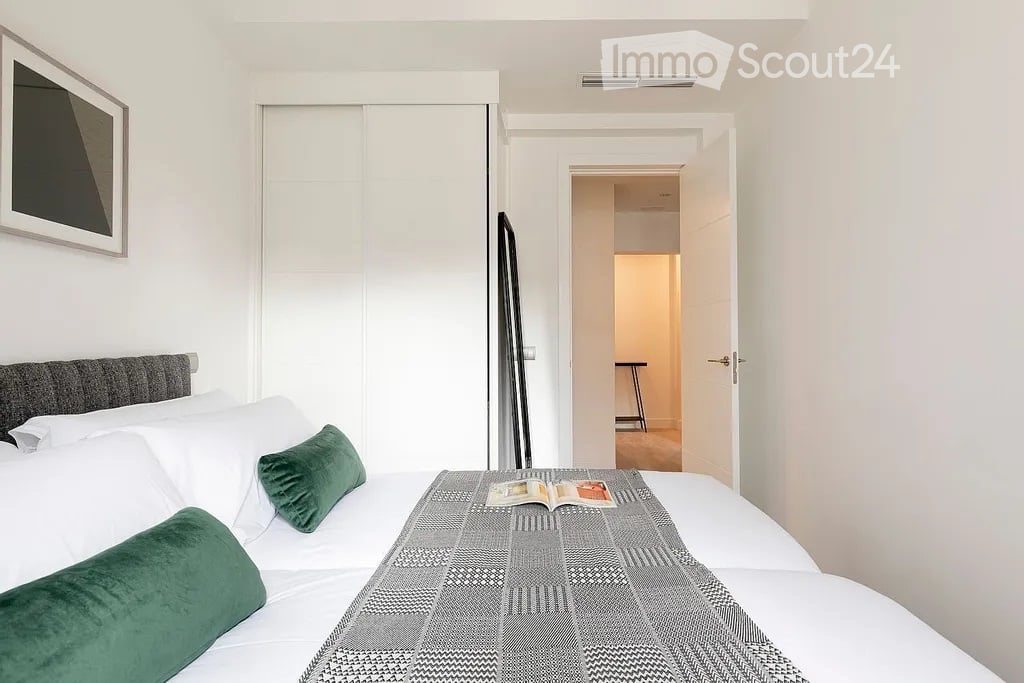Modern bedroom with a neatly made bed, green throw pillows, a grey headboard, and a picture frame on the wall. An open door leads to another room with a visible stool.