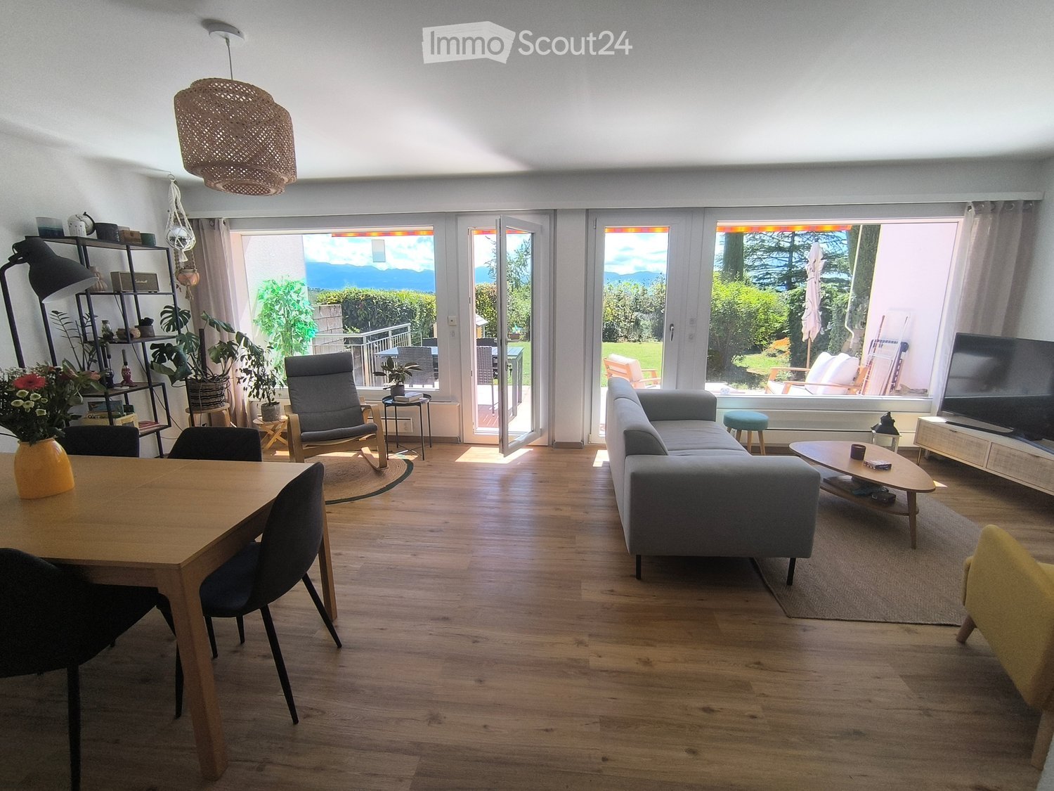 Bright living and dining area with wooden flooring, large windows offering a scenic view, a grey sofa, a coffee table, a dining table with chairs, a shelf with plants and decor, and a television on a stand.