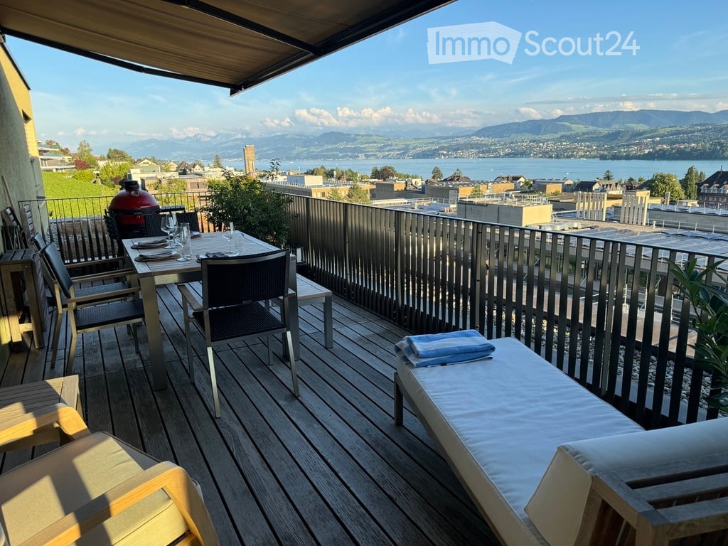 Balcony with wooden decking, outdoor dining set, sunbed, and grill, overlooking a scenic lake and mountain view.