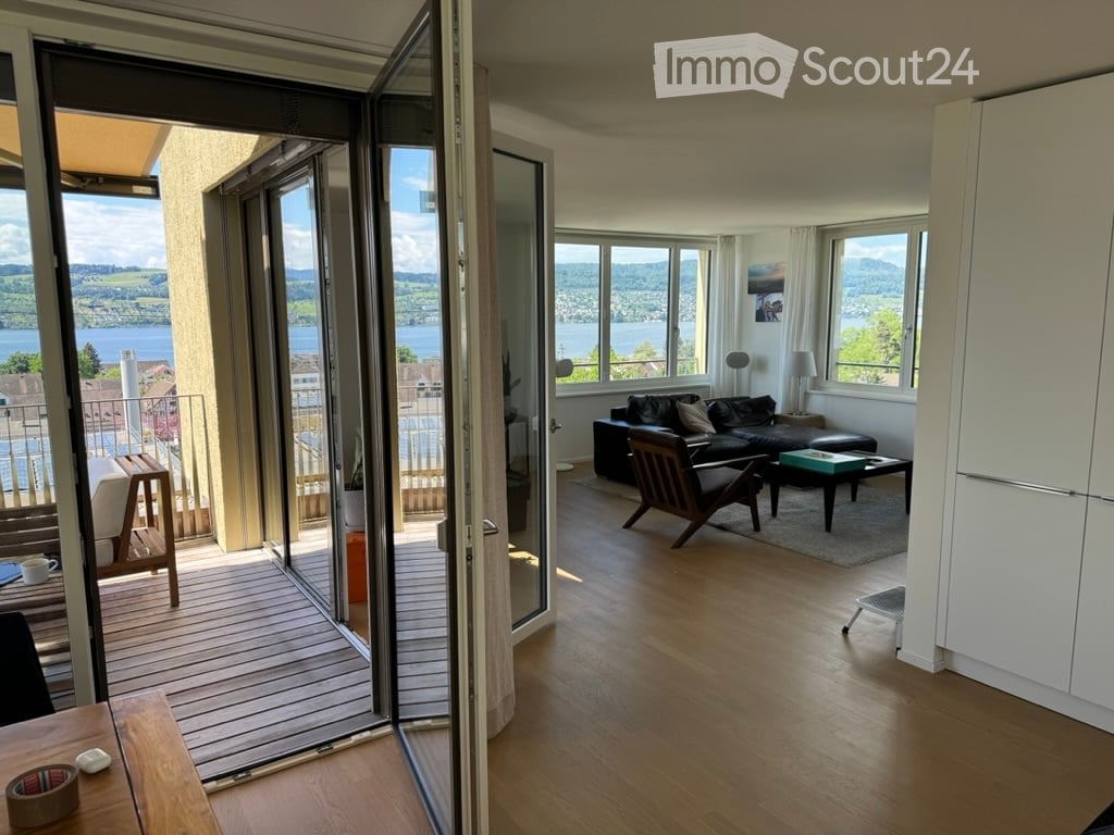 Bright living room with large windows and a balcony overlooking a scenic lake view. Features include a black leather sofa, a wooden coffee table, a wooden chair, and a small table with a cup on it.