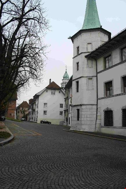 A quiet cobblestone street lined with historic white buildings, including a prominent tower with a green spire and a smaller church tower in the background.