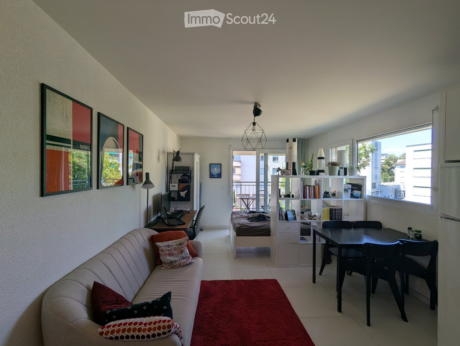 A modern, open-concept living and sleeping area with a sofa, a small bed, a dining table, a laptop on a desk, and shelves with books and decorative items.