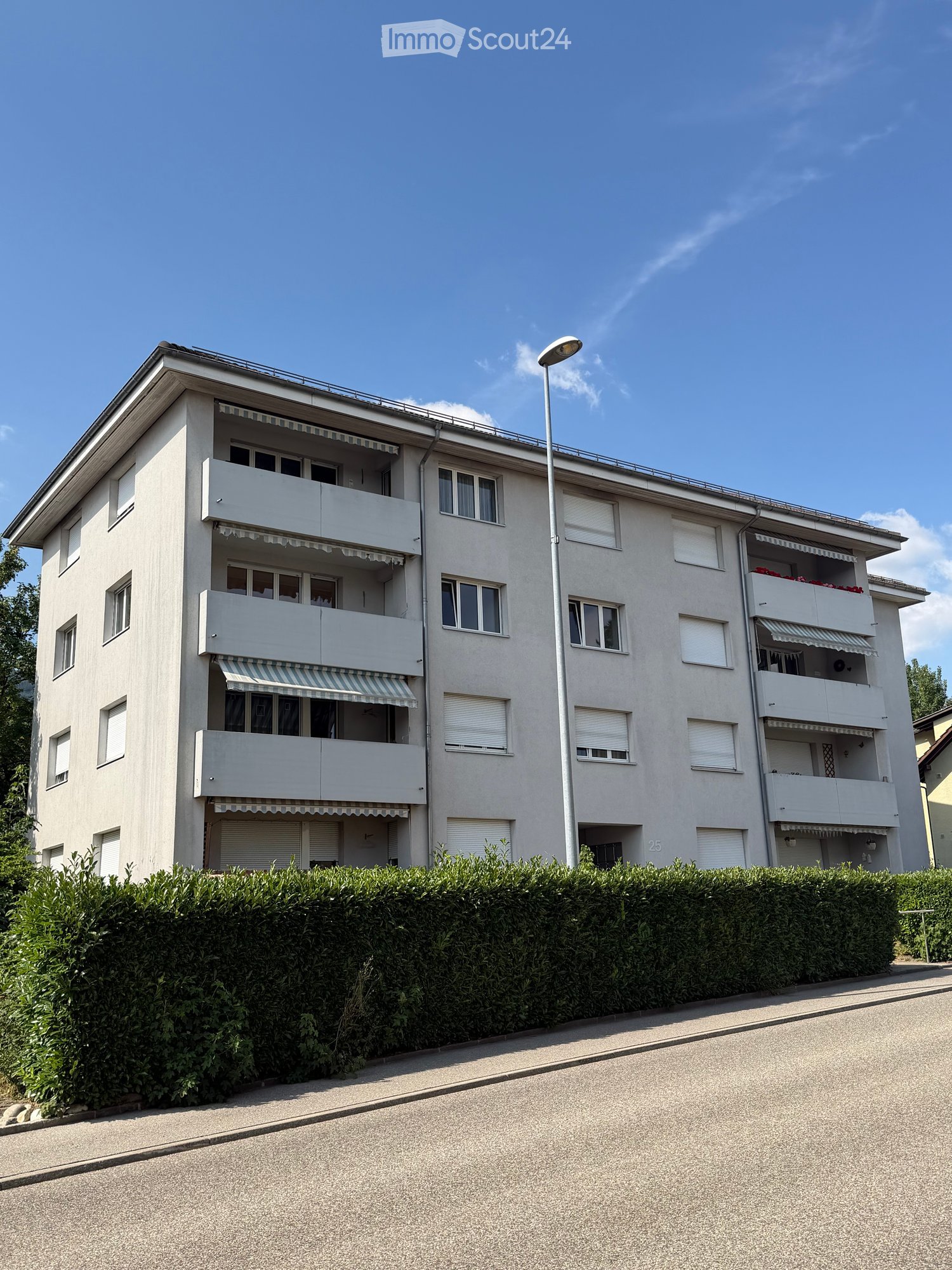 A modern multi-story apartment building with balconies, large windows, and striped awnings. The building is surrounded by a neatly trimmed hedge and a streetlight is visible on the sidewalk.