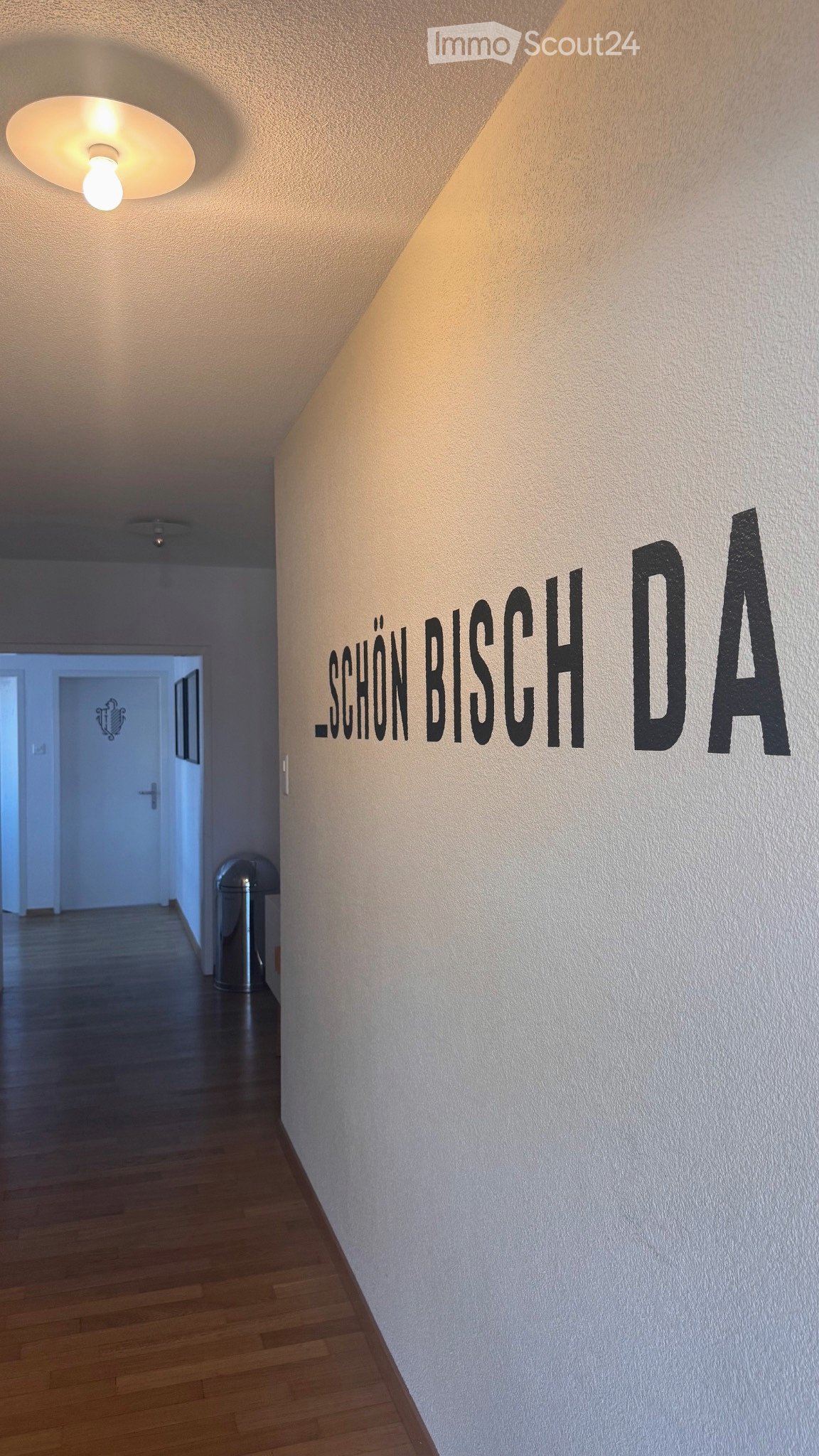 A well-lit hallway with wooden flooring, white walls, and a decorative sign reading "SCHON BISCH DA" on the right wall. A trash bin is visible near the left side, and a door is seen at the far end of the corridor.
