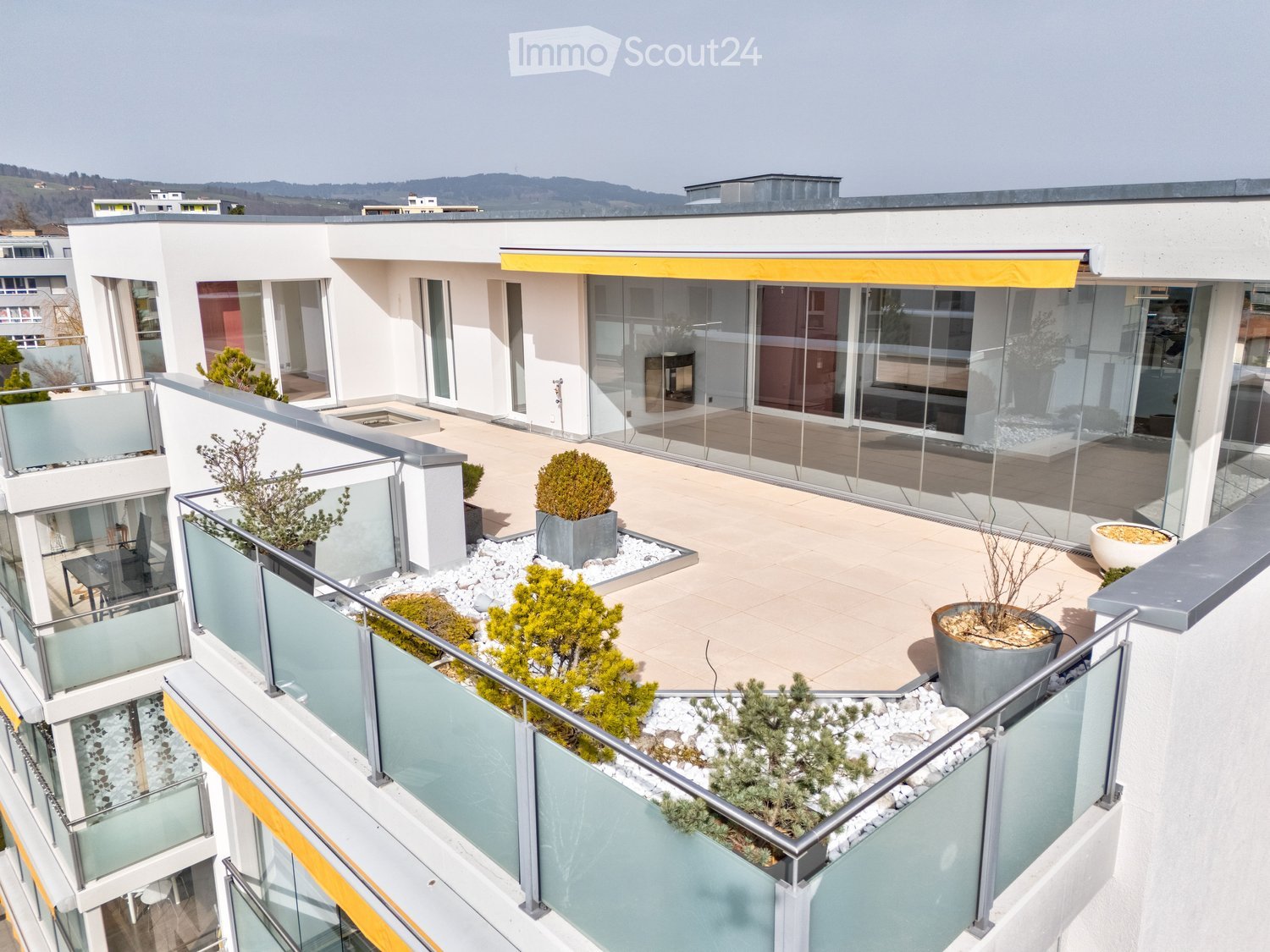Modern rooftop terrace with glass railings, potted plants, and a view of the surrounding area.